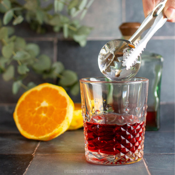 Glass with red liquid, orange, and tongs on a kitchen counter. Clear ice sphere maker, ice ball press kit by pressice barware.