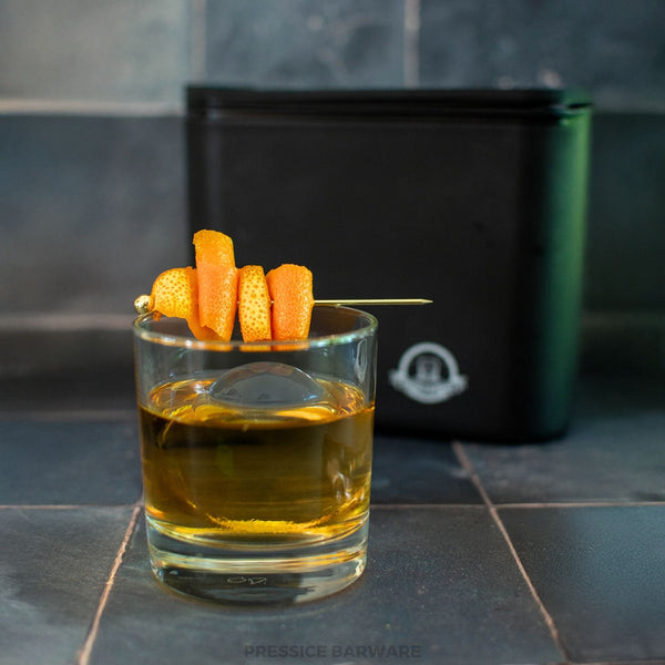 Glass of amber liquid with a skewer of orange peel on a dark tiled surface, featuring Pressice Barware branding.