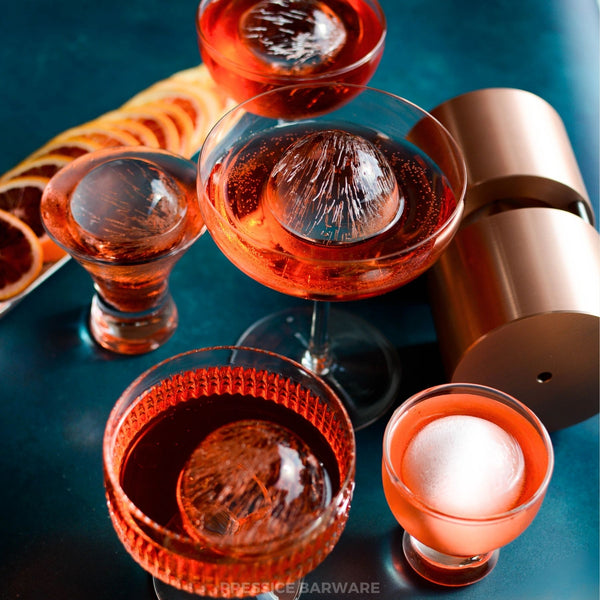 Cocktail glasses with red liquid and ice cubes on a blue surface. Clear ice spheres and ice ball press by Pressice Barware