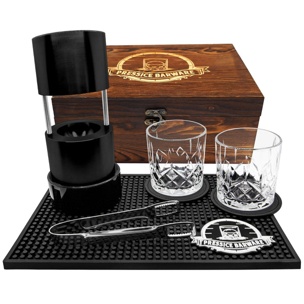 Barware set with two glasses, a black container, and tools on a black mat with a wooden box labeled 'Pressie Barware'.